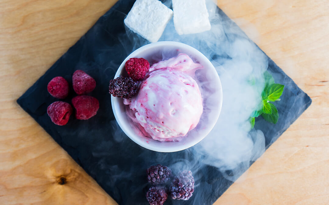 Top view of a cup of Ice Cream next to liquid nitrogen, fruit, and herbs. All sitting on a black stone slab.