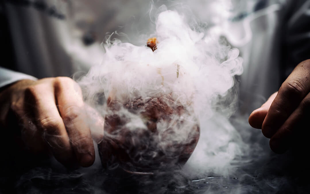 A chef's hands around a glass with a drink that has nitrogen smoke coming off.