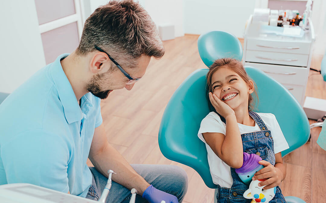 A dentist sitting next to a smiling child holding some toys and touching her cheek.