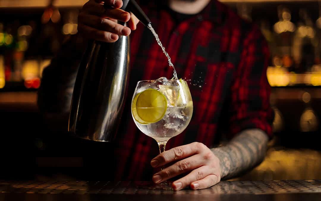 Bartender adding tonic water to the to the cocktail glass full of ice cubes with lime slices on the steel bar counter