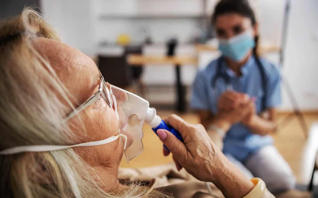 Woman sitting at home and taking oxygen from a respirator while nurse with protective mask on sitting next to her and holding her hand during coronavirus.